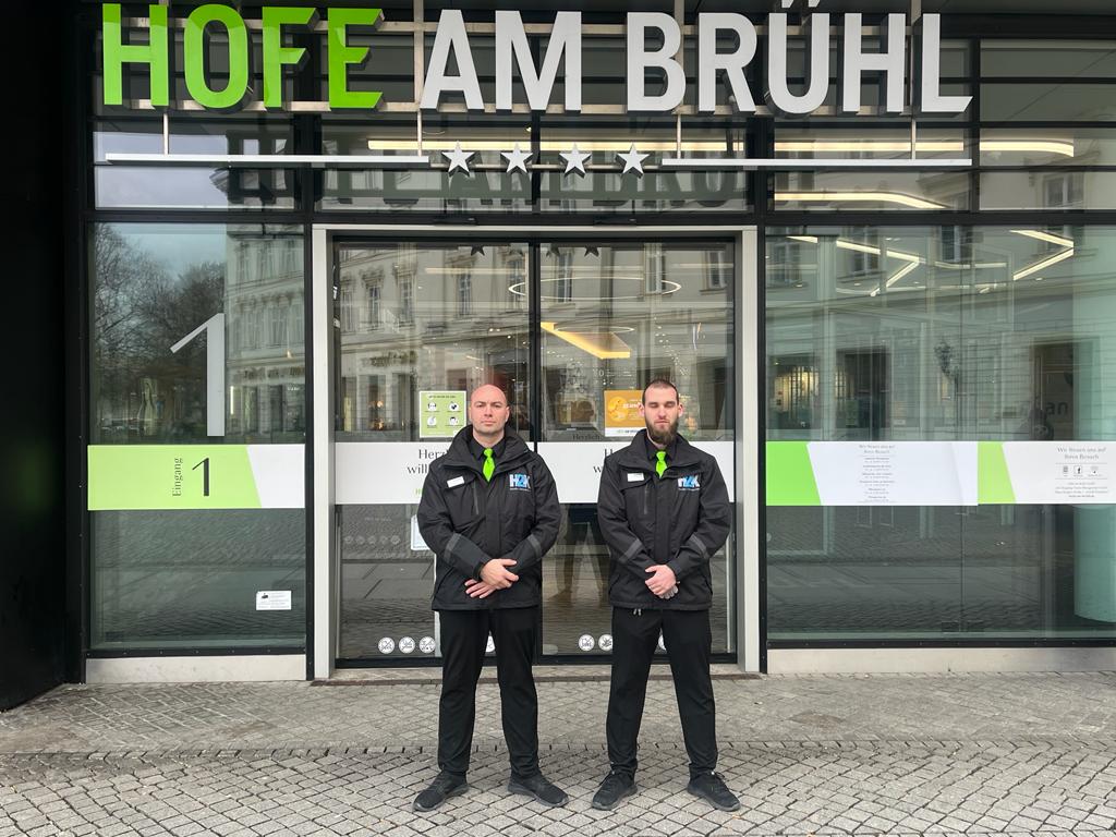 Zwei H2K-Mitarbeiter in H2K-Arbeitsjacken stehen vor dem Hofe am Brühl.