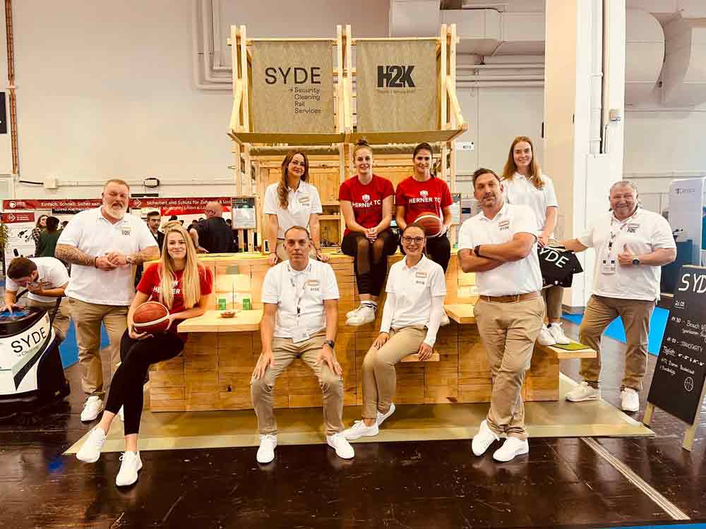Mehrere Mitarbeiter von H2K und Syde in einheitlicher Arbeitskleidung, zwei von ihnen halten Basketbälle. Hinter ihnen steht ein Stand mit den Firmenlogos.