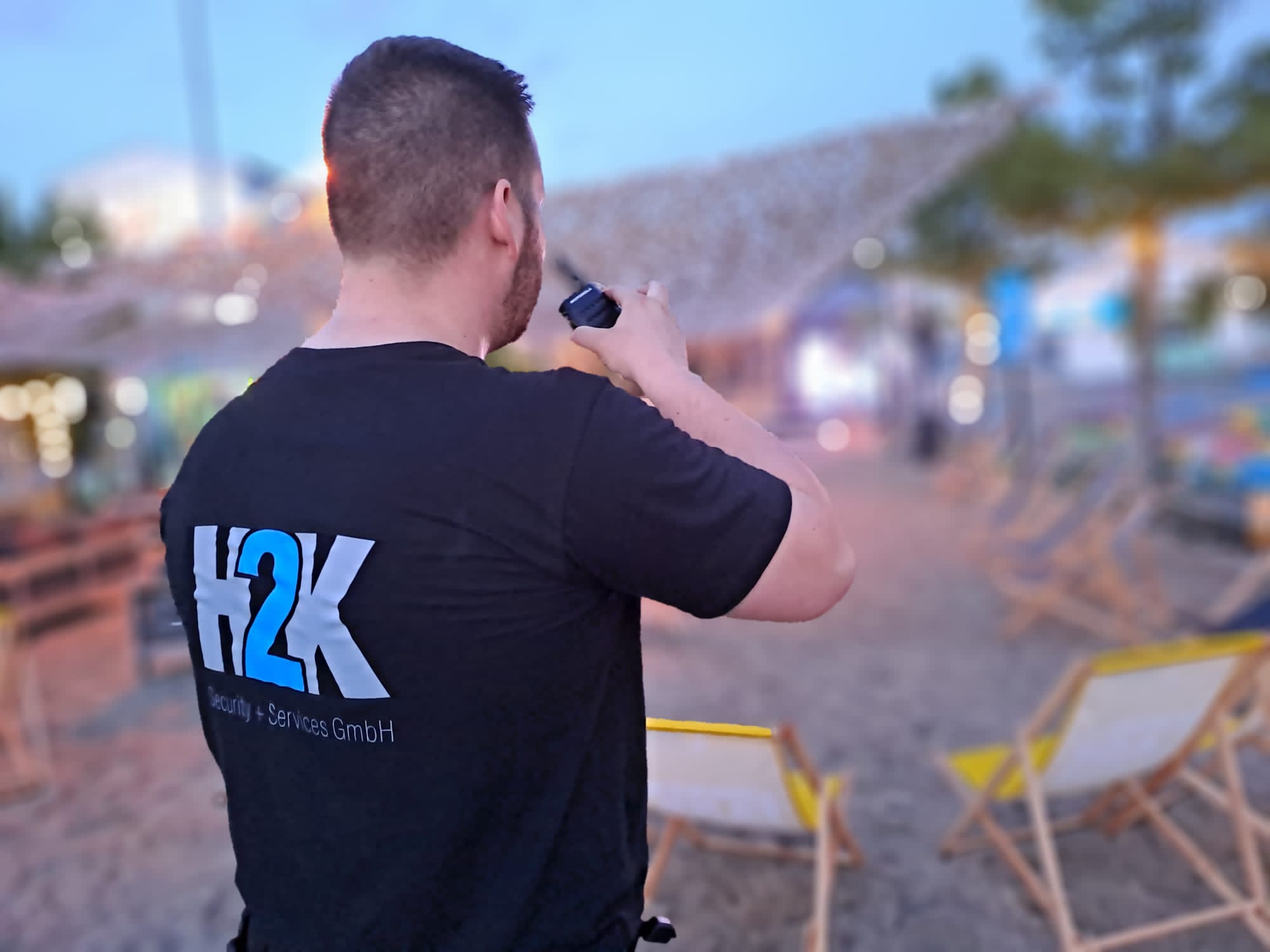in H2K-Mitarbeiter in H2K-Arbeitskleidung mit einem Walkie-Talkie in der Hand. Im Hintergrund ist ein Strandbereich.